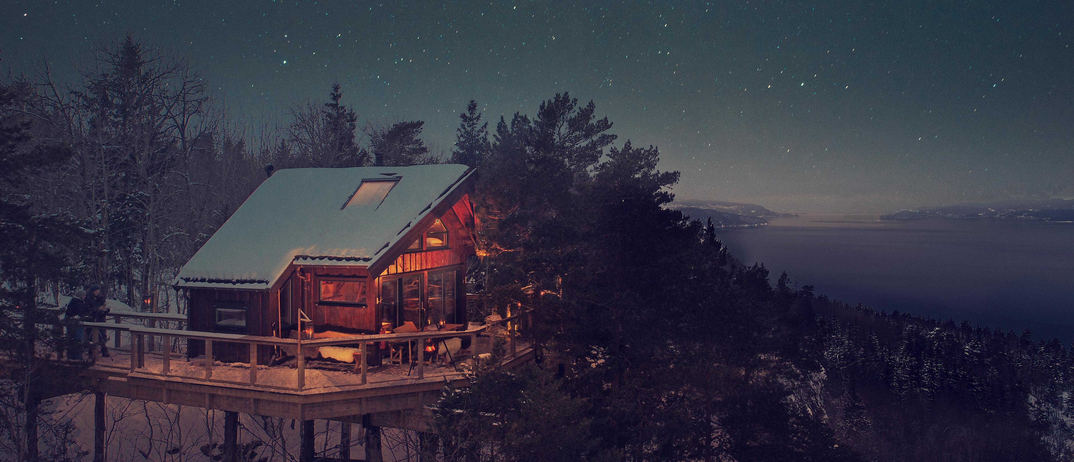 Drone photo with the Oslofjord in the background and an illuminated treetop cabin with many trees around.