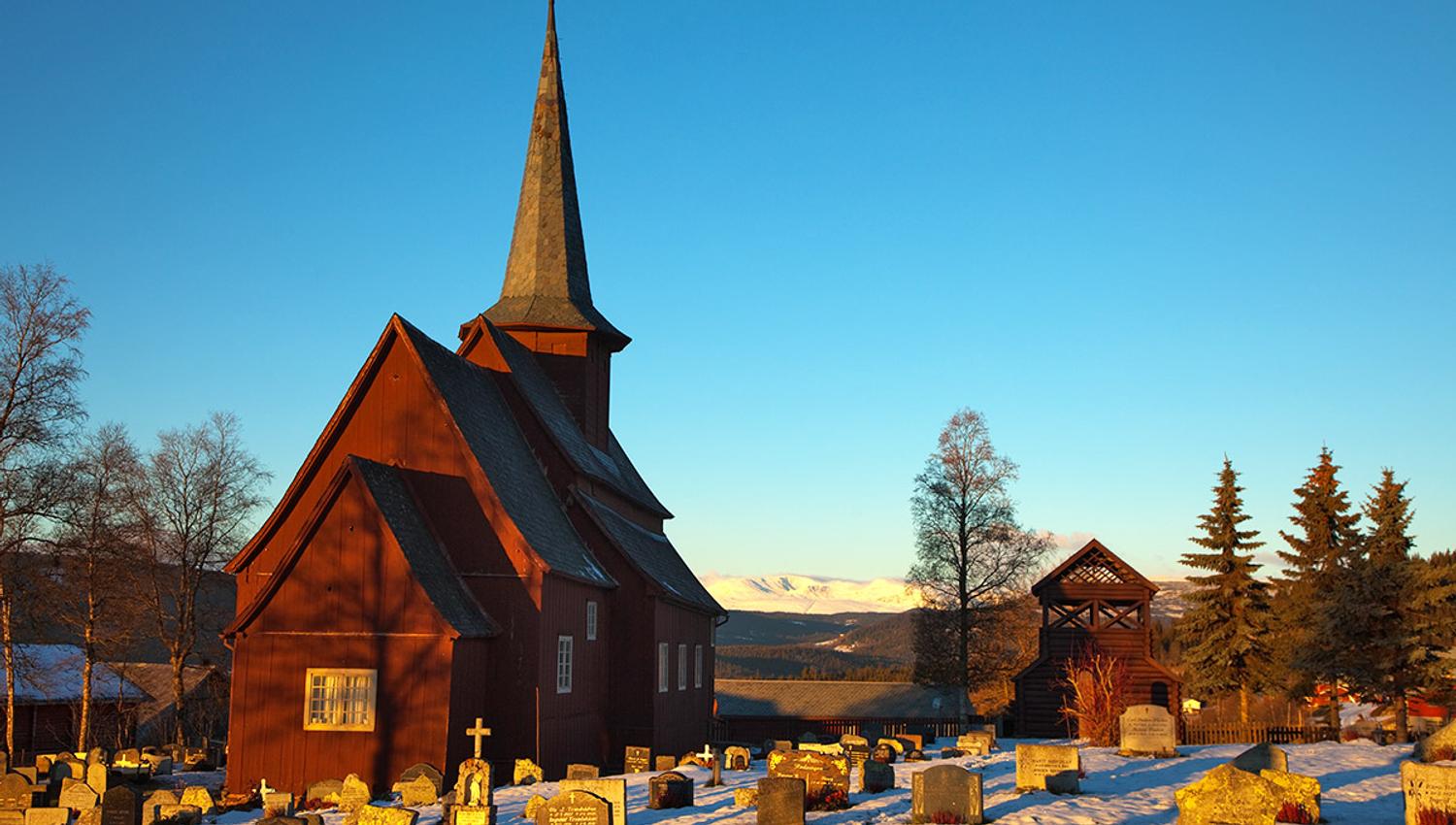 A stave church in a snowy church yard lit by winter sunshine.
