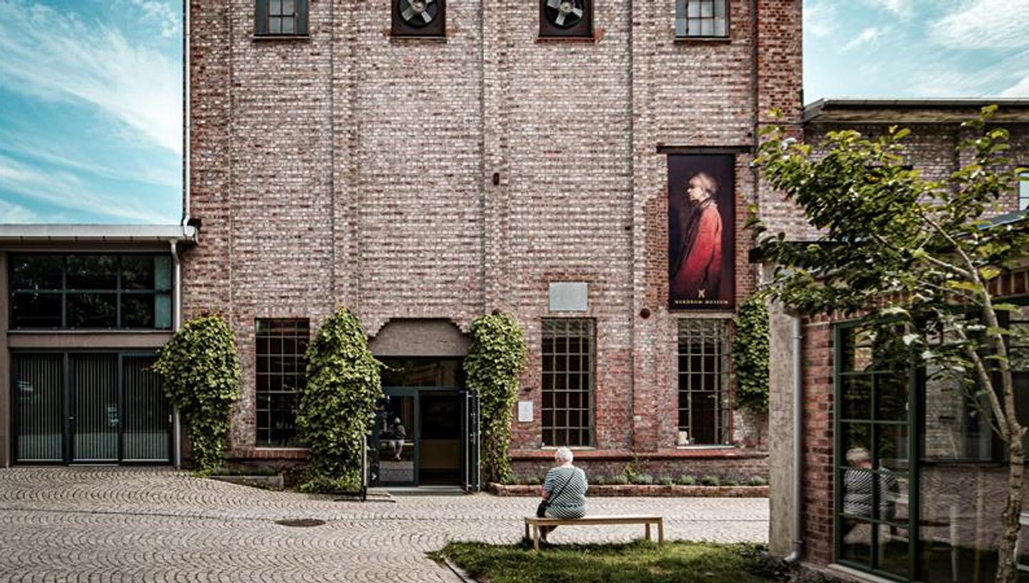 Nærbilde av museumsbygget. En person sitter på en benk.