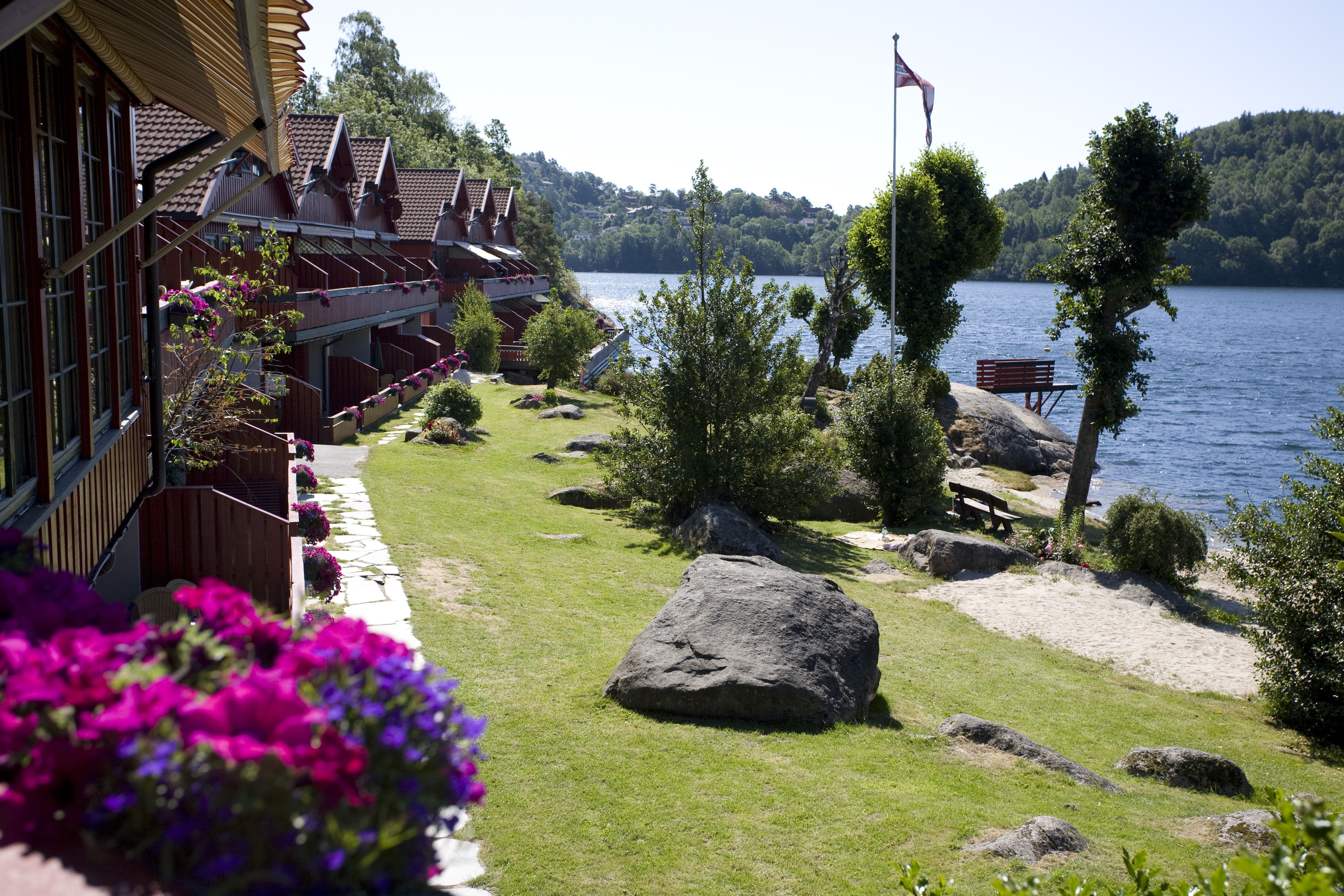 feriehus med rosalilla blomster i blomsterkasse på alle terrassene på venstre side med nyklipt hage og vann på høyre side. Norsk vimpel på flaggstang. skyfri himmel.