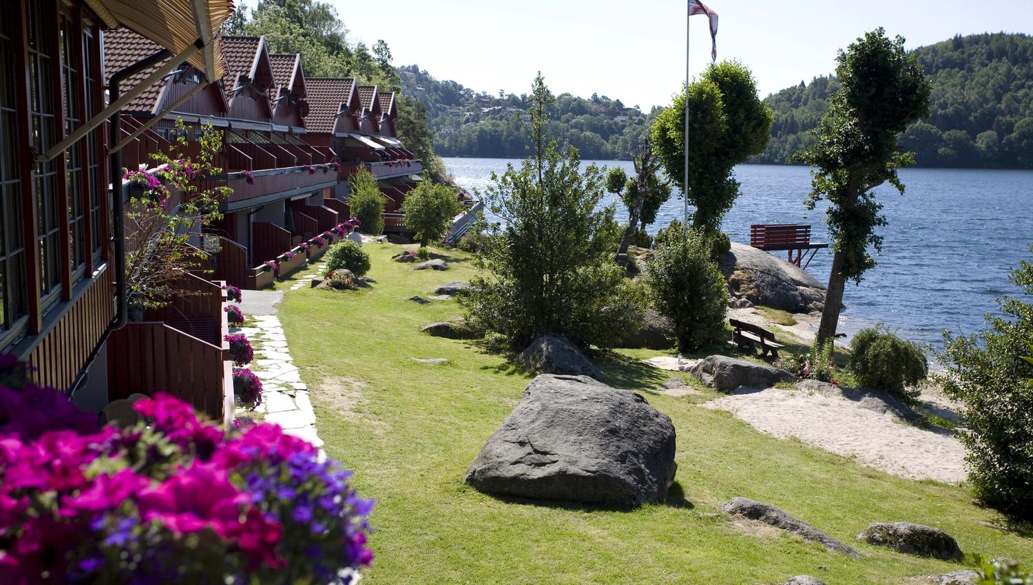 feriehus med rosalilla blomster i blomsterkasse på alle terrassene på venstre side med nyklipt hage og vann på høyre side. Norsk vimpel på flaggstang. skyfri himmel.