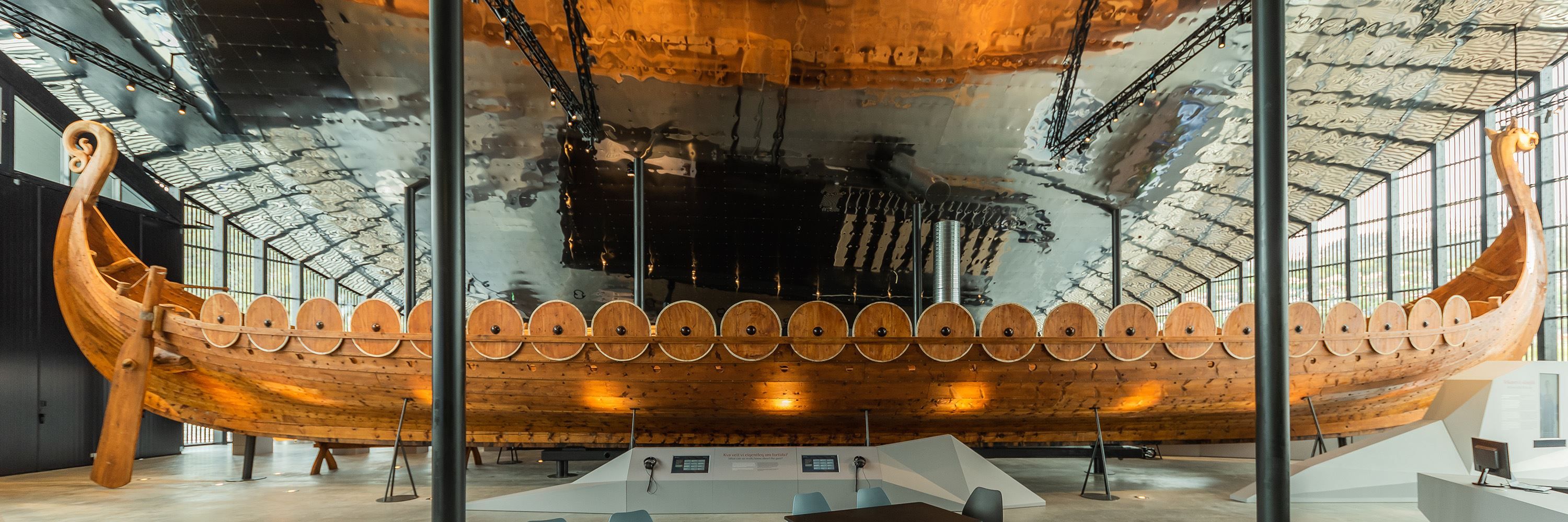 Det 30 meter lange Myklebustskipet utstilt inne på Sagastad Vikingsenter - Eit stort vikingskip i tre står utstilt innandørs i ein museumsal med metalltak, vindauge og lyspunkter..
