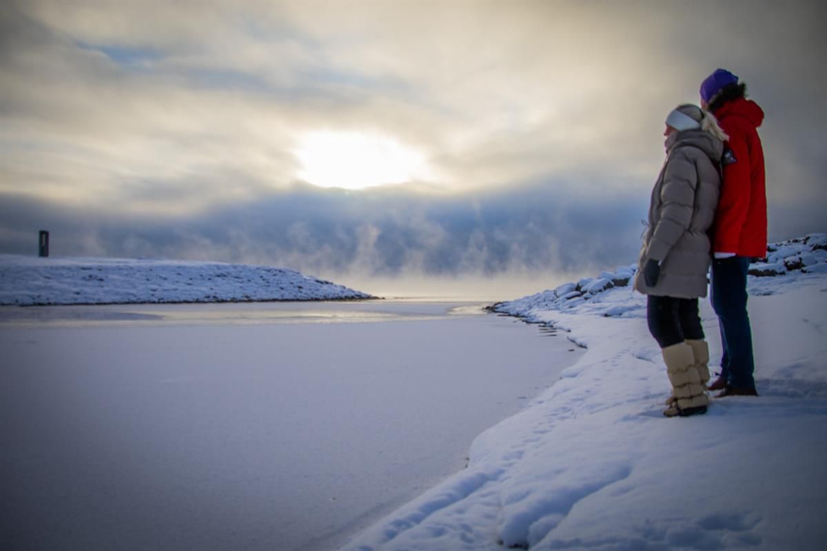 Personer står ved et snødekt vannkantområde med lav sol og skyer over horisonten.