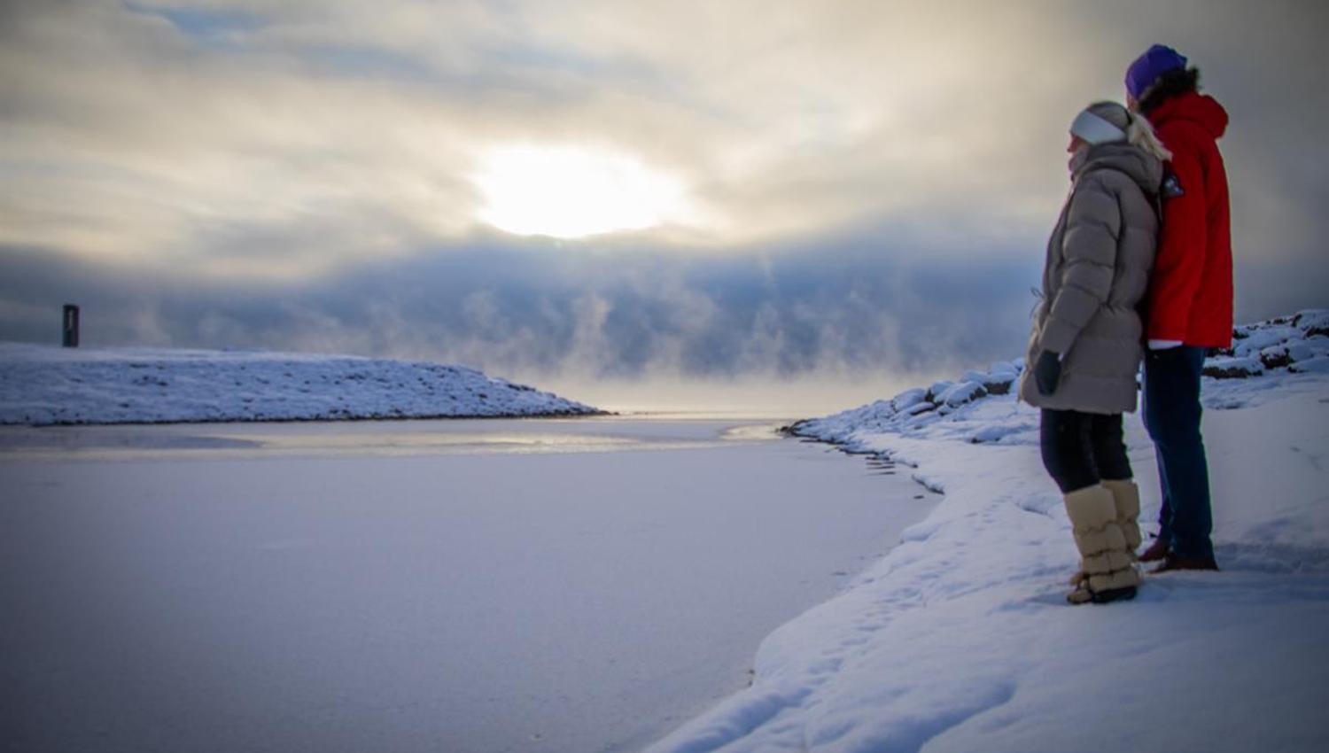 Personer står ved et snødekt vannkantområde med lav sol og skyer over horisonten.