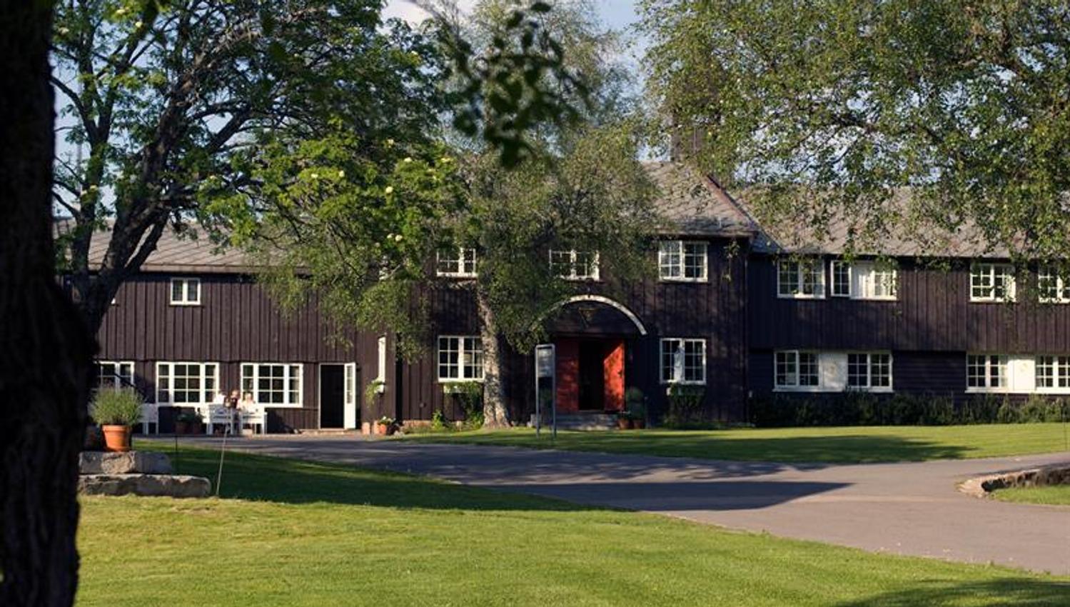 Lyseby fasade. Brunt trebygg med to etasjer og buet inngang. Gressplen og trær foran. Foto