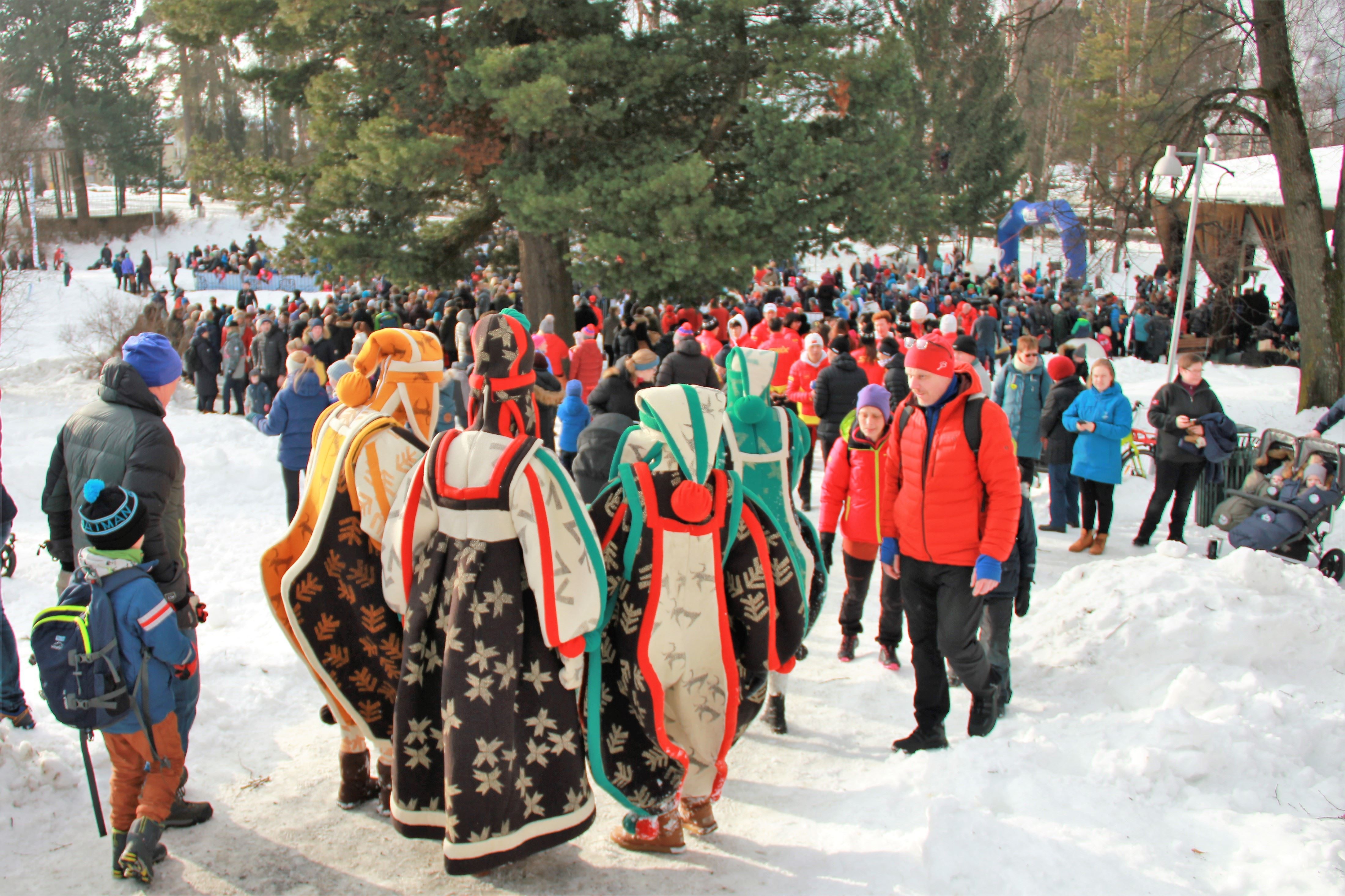 Stor folkemengde i snødekt park; flere går på sti, og tre personer i fargerike kapper sees bakfra..