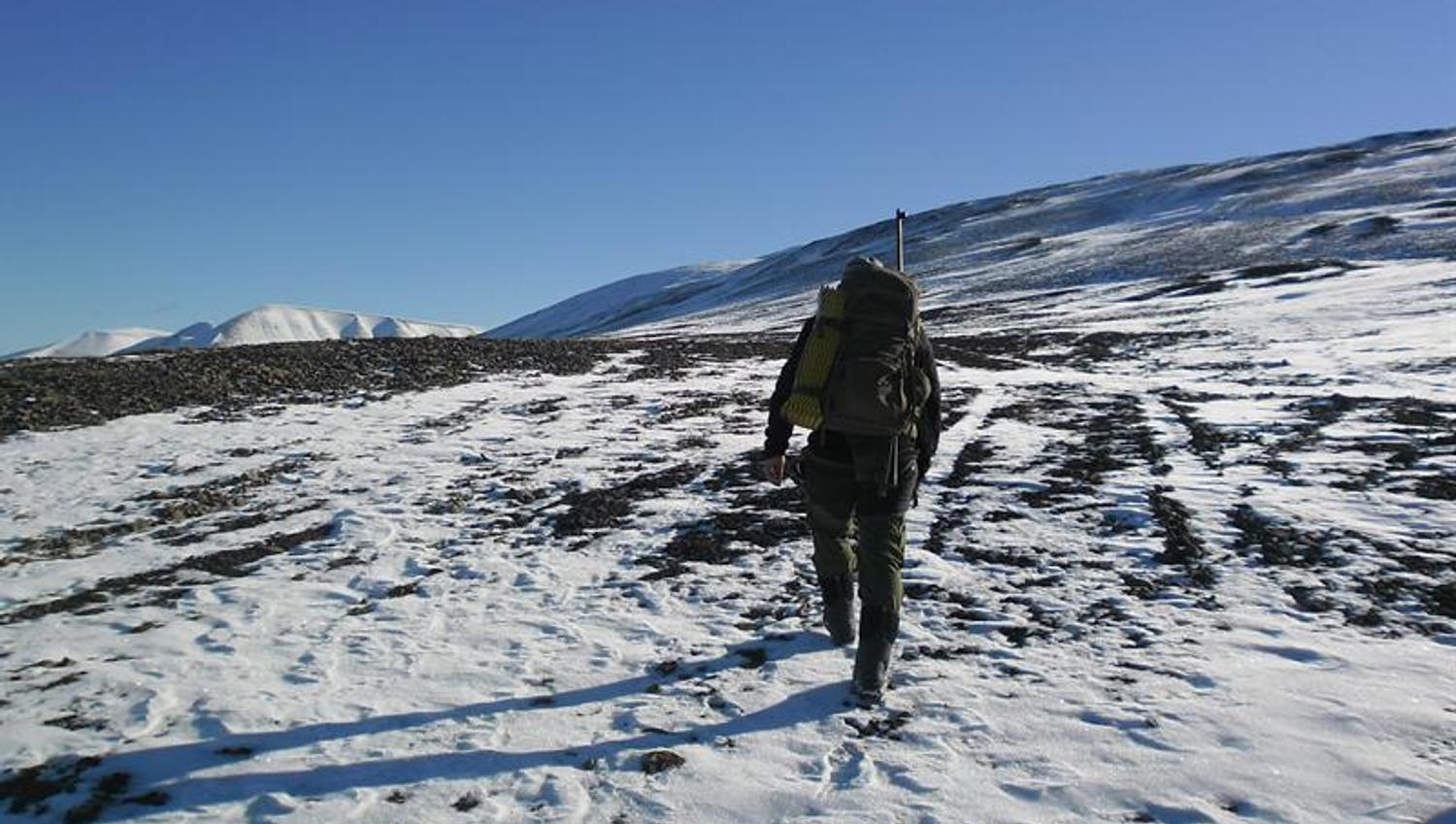 En guide med rifle og ryggsekk på ryggen som går gjennom et fjellandskap dekket i snø