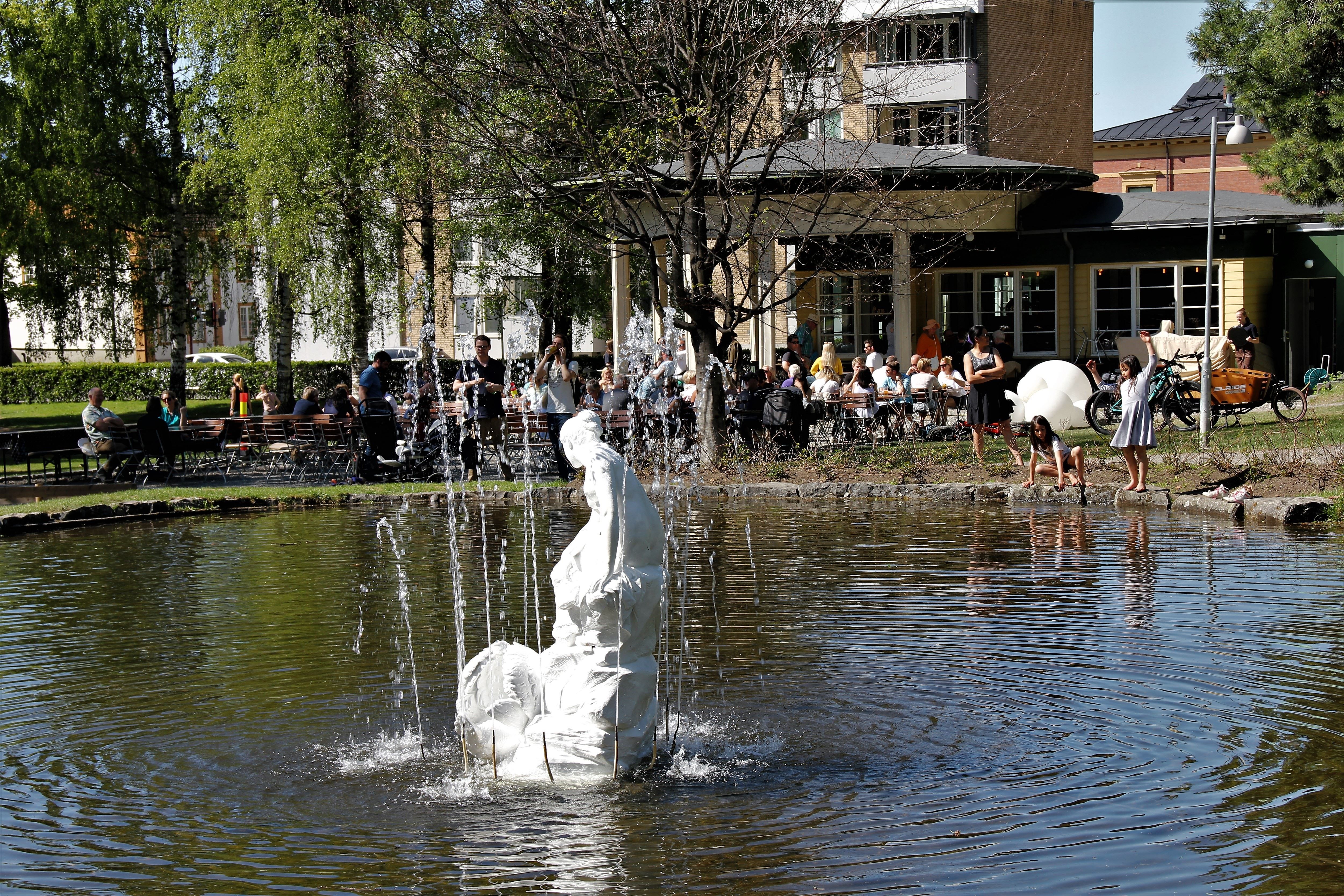 Hvit skulptur i en fontene i en dam, med kaféservering og folk under trær ved bygninger i bakgrunnen..