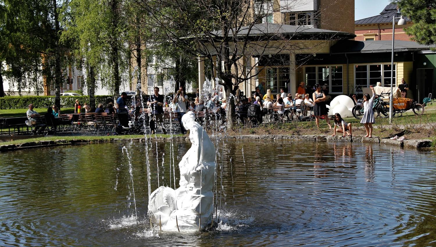 Hvit skulptur i en fontene i en dam, med kaféservering og folk under trær ved bygninger i bakgrunnen..