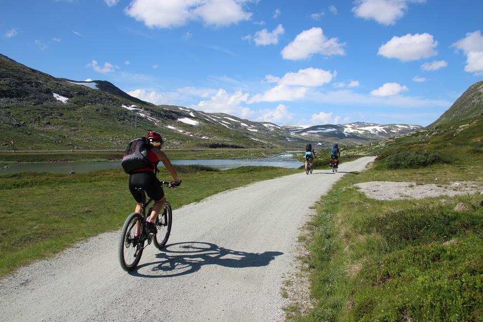 Syklistar nyt turen på Rallarvegen langs stille fjellvatn under blå himmel.