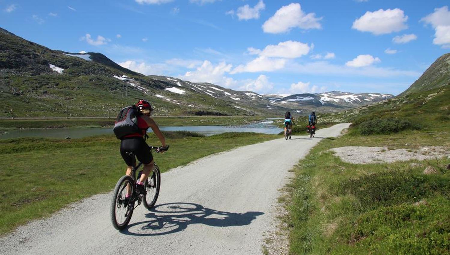 Syklistar nyt turen på Rallarvegen langs stille fjellvatn under blå himmel.