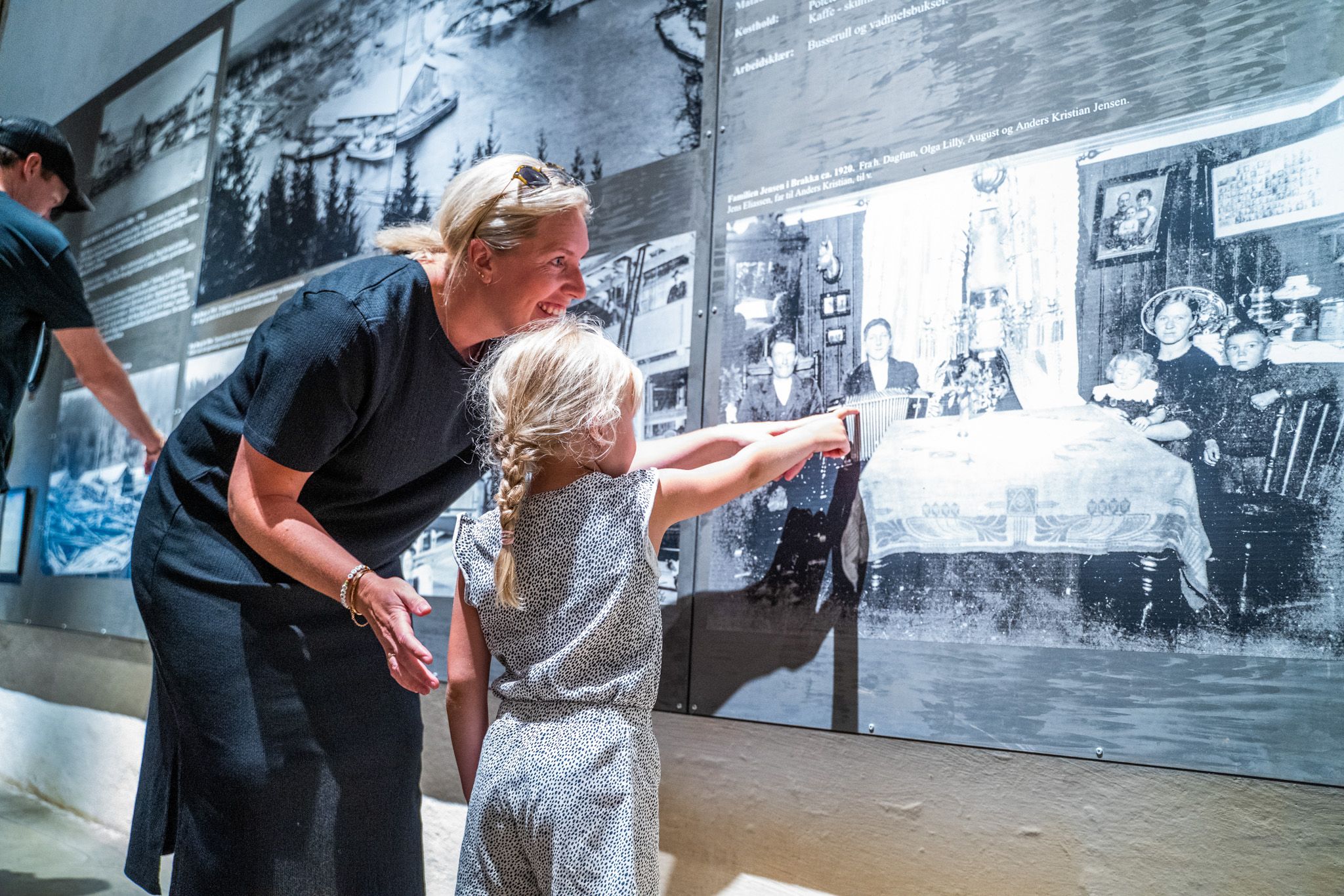 Bildet viser en kvinne og en liten jente som ser på store svart-hvite bilder på en museumsvegg. Jenta peker på et av bildene, og kvinnen smiler mens hun ser på det sammen med henne. Stemningen er varm og nysgjerrig.