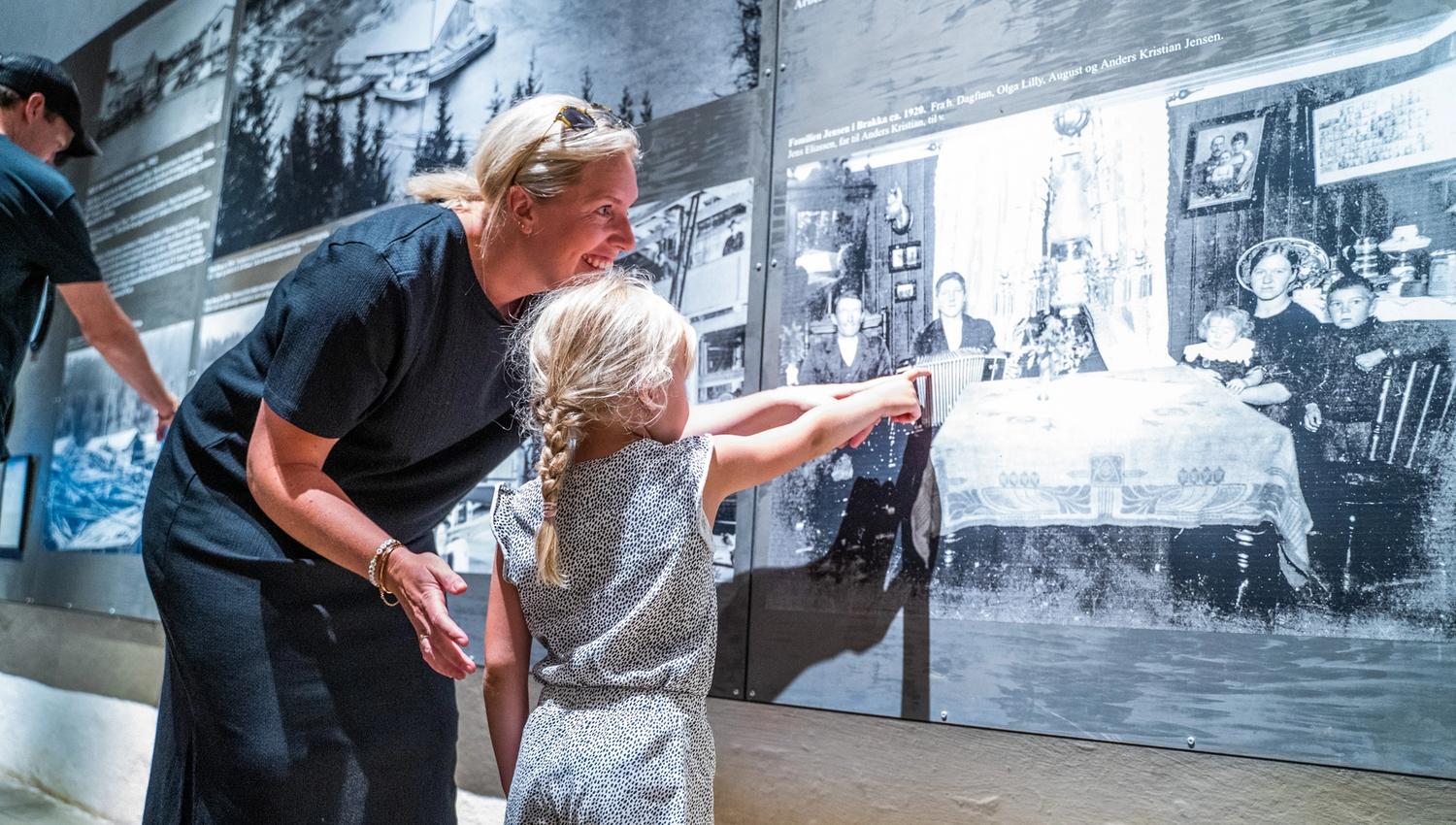 Bildet viser en kvinne og en liten jente som ser på store svart-hvite bilder på en museumsvegg. Jenta peker på et av bildene, og kvinnen smiler mens hun ser på det sammen med henne. Stemningen er varm og nysgjerrig.