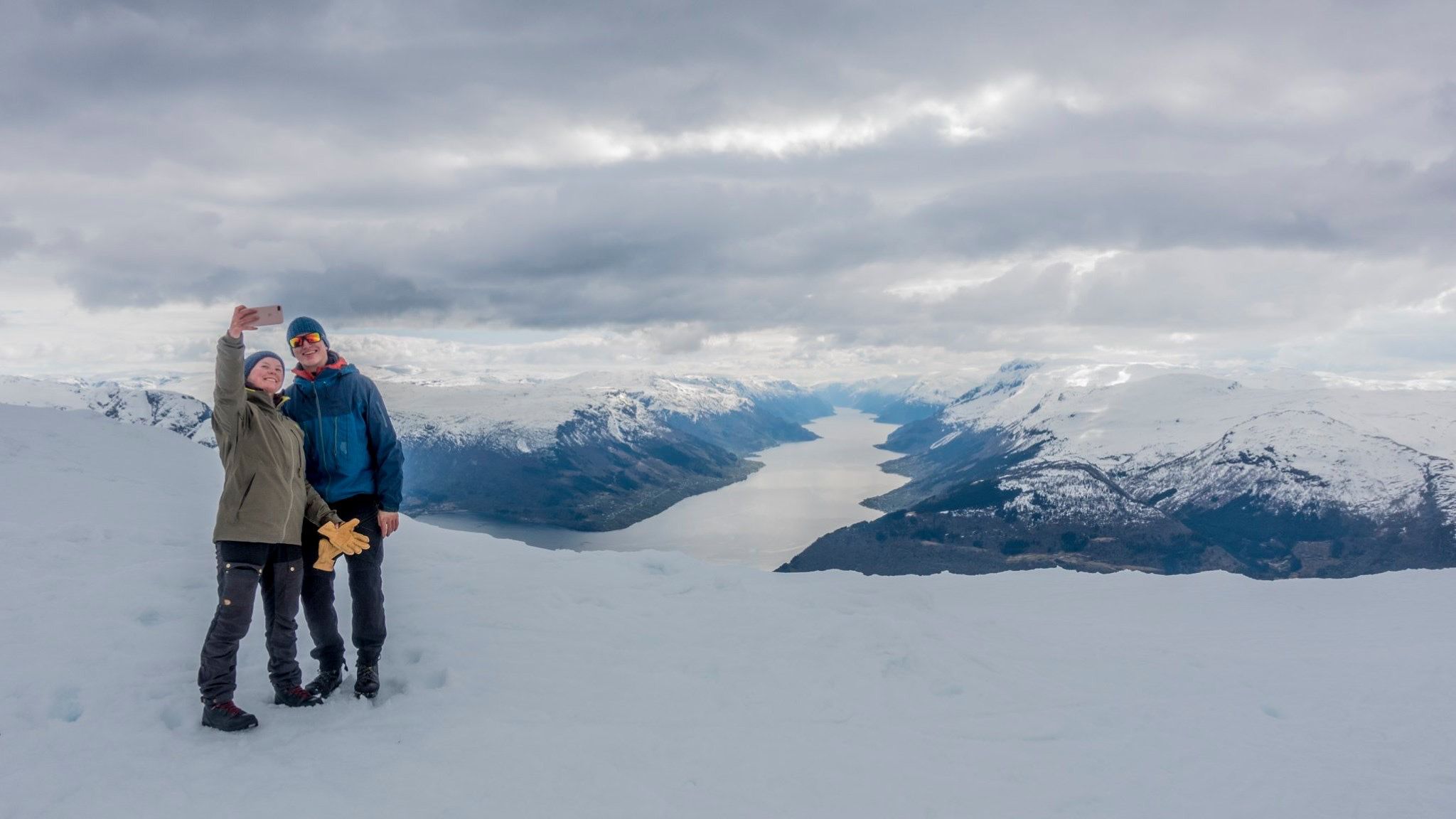 Couple taking a selfie while snowshoeing with dramatic fjord scenery behind them.
