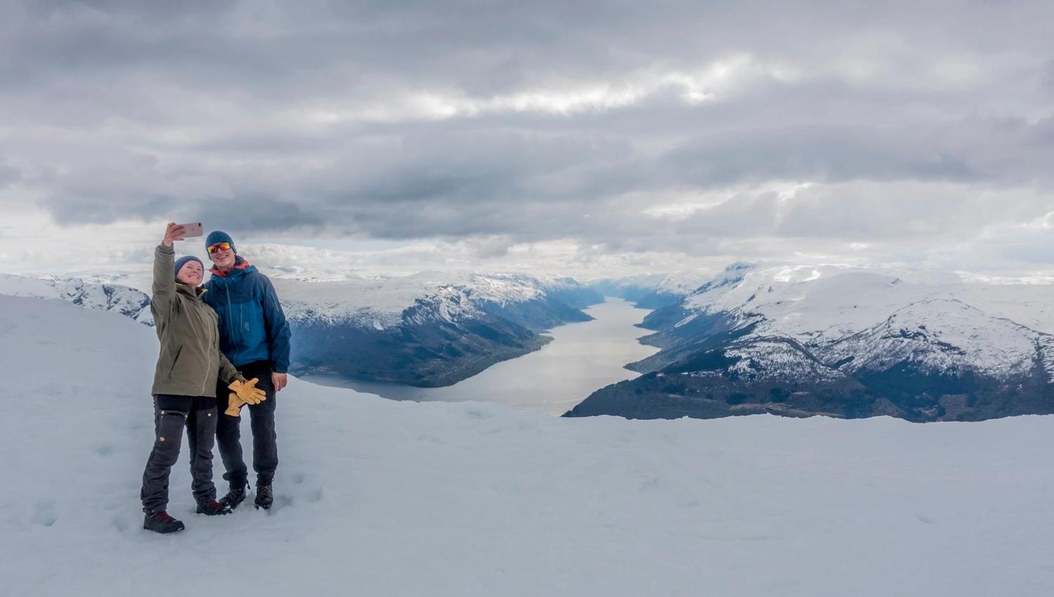 Couple taking a selfie while snowshoeing with dramatic fjord scenery behind them.