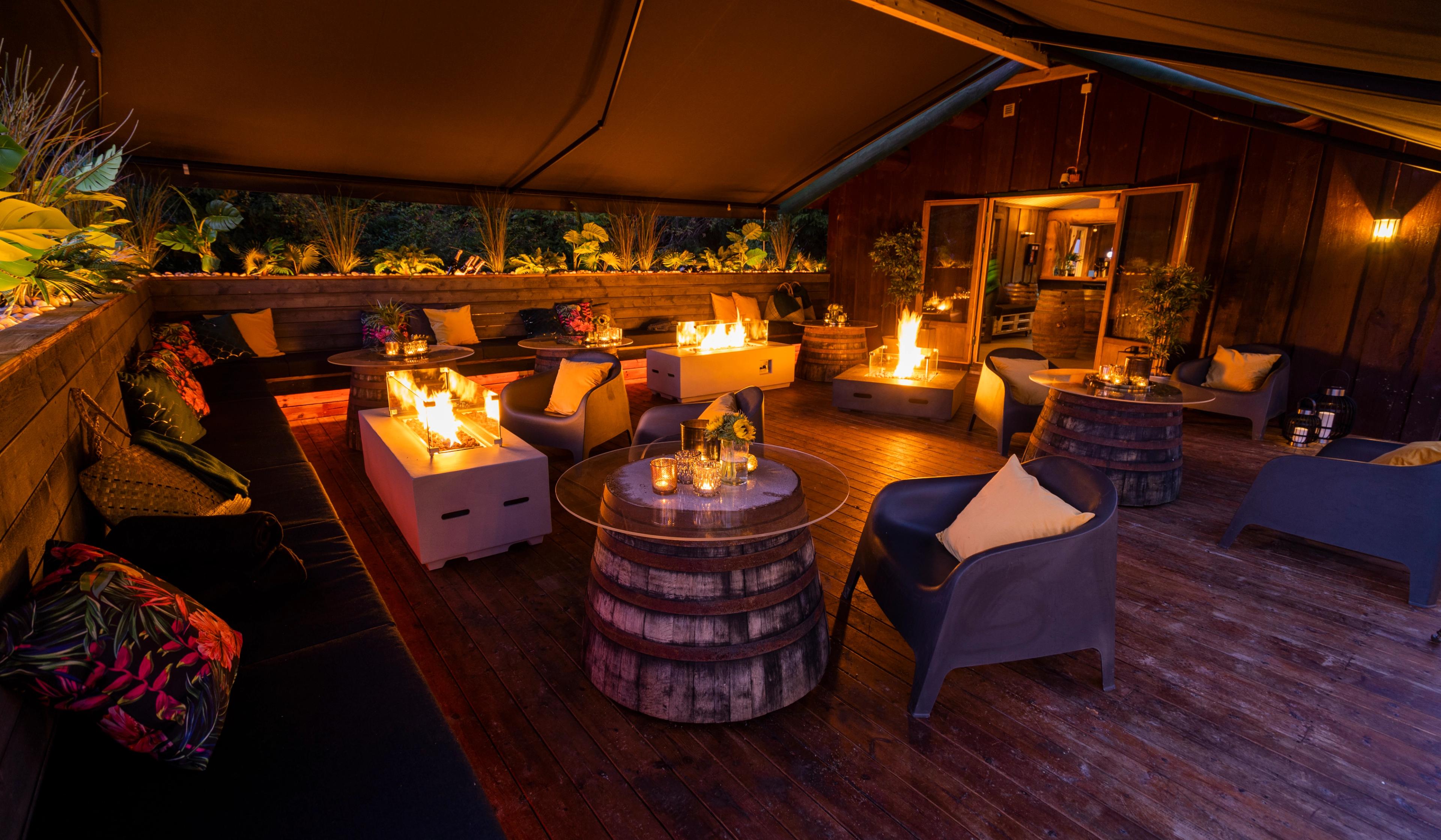 Uteområde under tak med komfortable sitteplasser, tønnebord og flere gasspeiser som gir varme og stemningsfull belysning. Området har trevegger og dekorative detaljer, omgitt av natur.