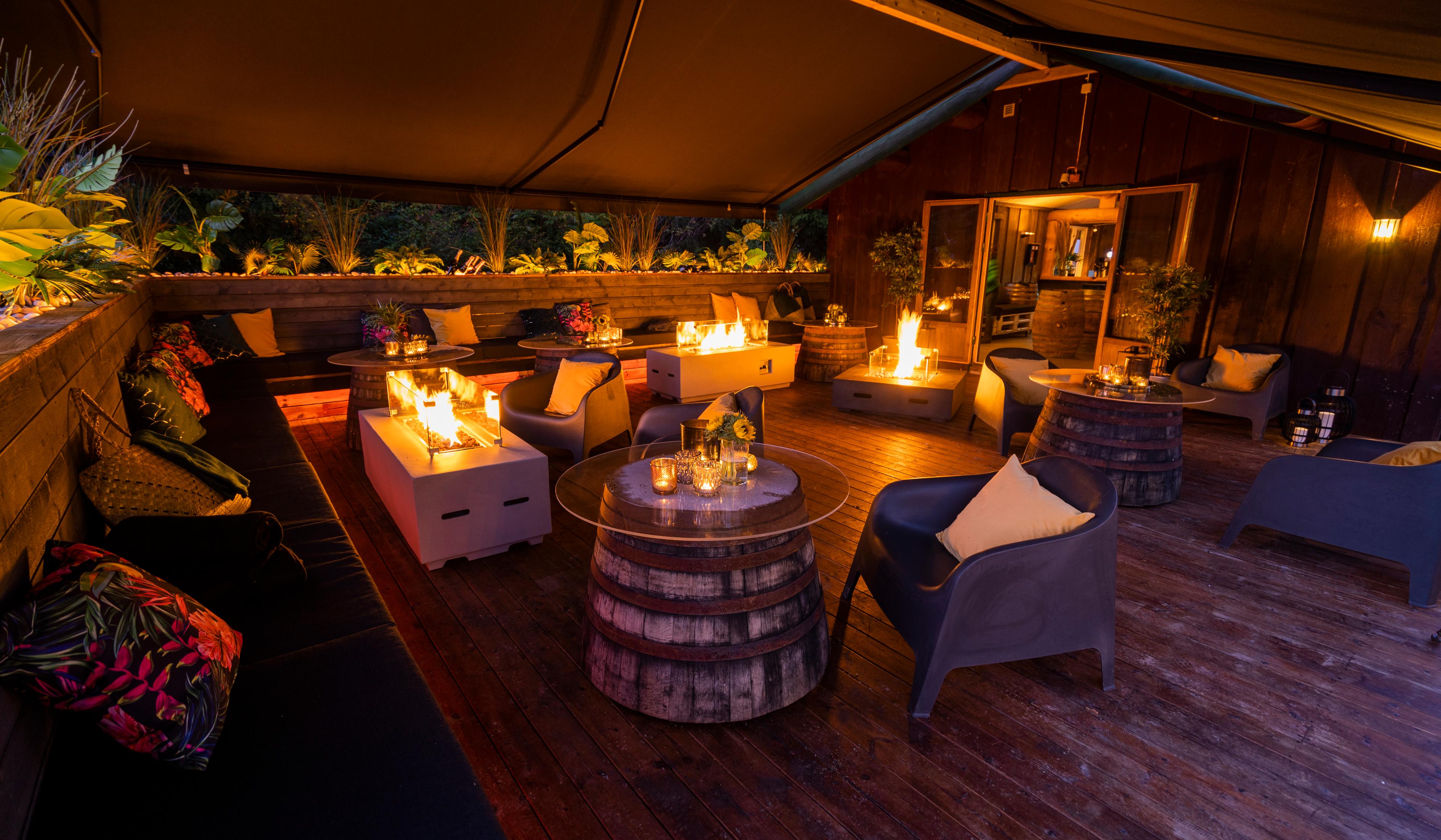 Uteområde under tak med komfortable sitteplasser, tønnebord og flere gasspeiser som gir varme og stemningsfull belysning. Området har trevegger og dekorative detaljer, omgitt av natur.