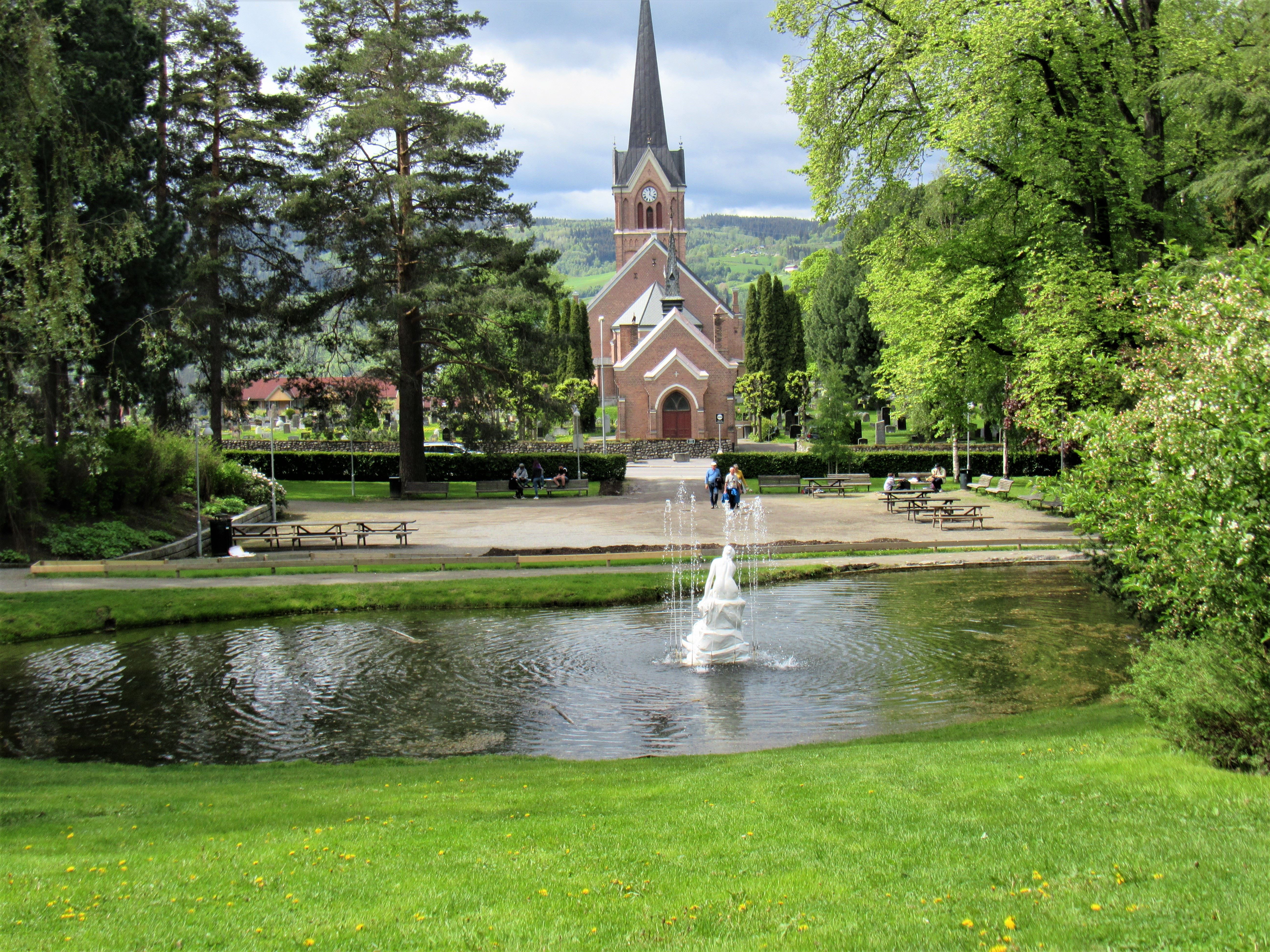 Mursteinskirke med høy spiss bak en grønn park; i forgrunnen er en dam med sprutende fontene og benker..