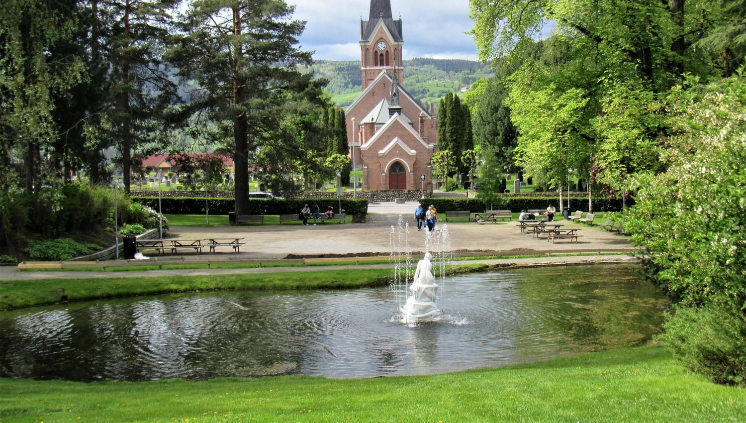 Mursteinskirke med høy spiss bak en grønn park; i forgrunnen er en dam med sprutende fontene og benker..