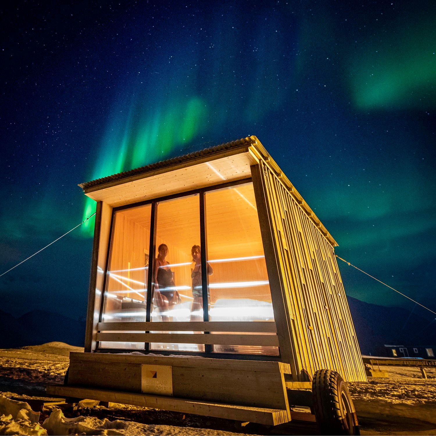 Two persons in the sauna at Isfjord Radio looking out through a window. Outside it's dark with stars and northern lights in the skies. 