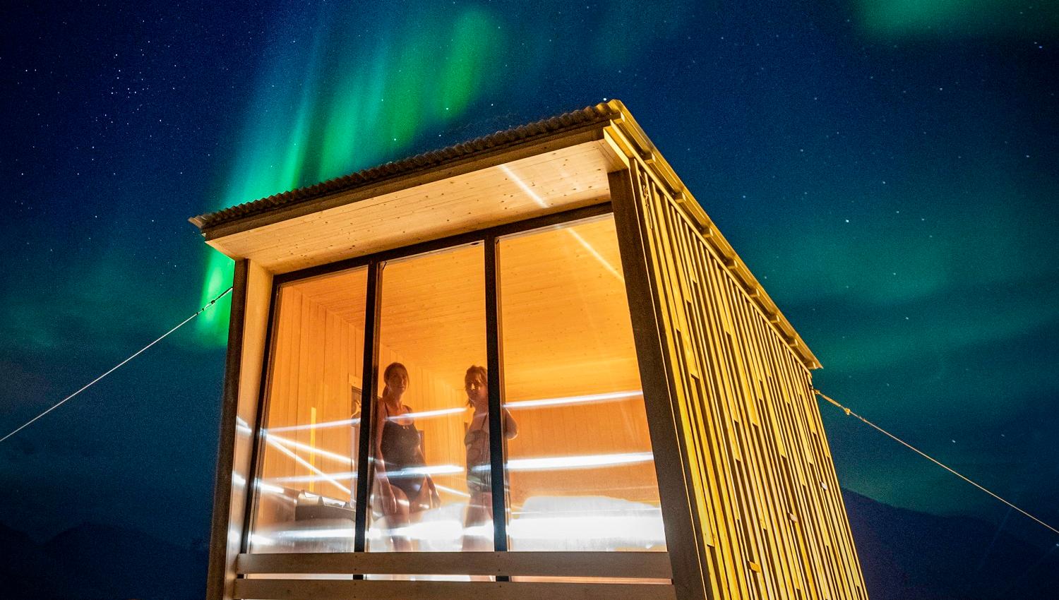 Two persons in the sauna at Isfjord Radio looking out through a window. Outside it's dark with stars and northern lights in the skies.