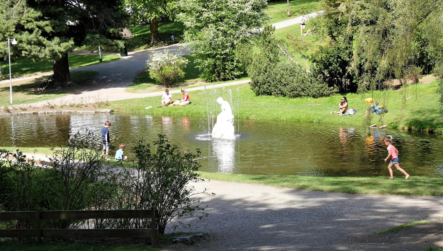 Folk slapper av på gresset rundt en parkdam med fontene, med trær, stier og sollys i bakgrunnen..