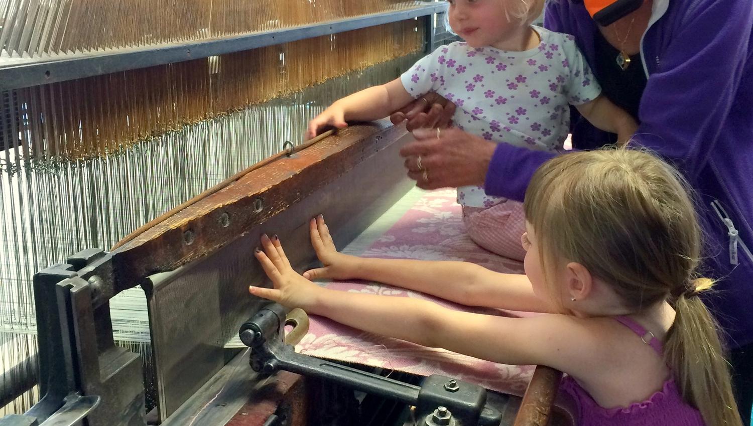Two children and an adult feeling and touching the weaving machine.