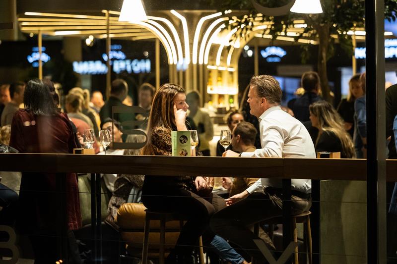 En kvinne og en mann sitter ved et bord og snakker sammen over et glass vin i et travelt og stemningsfullt restaurant- eller barområde.