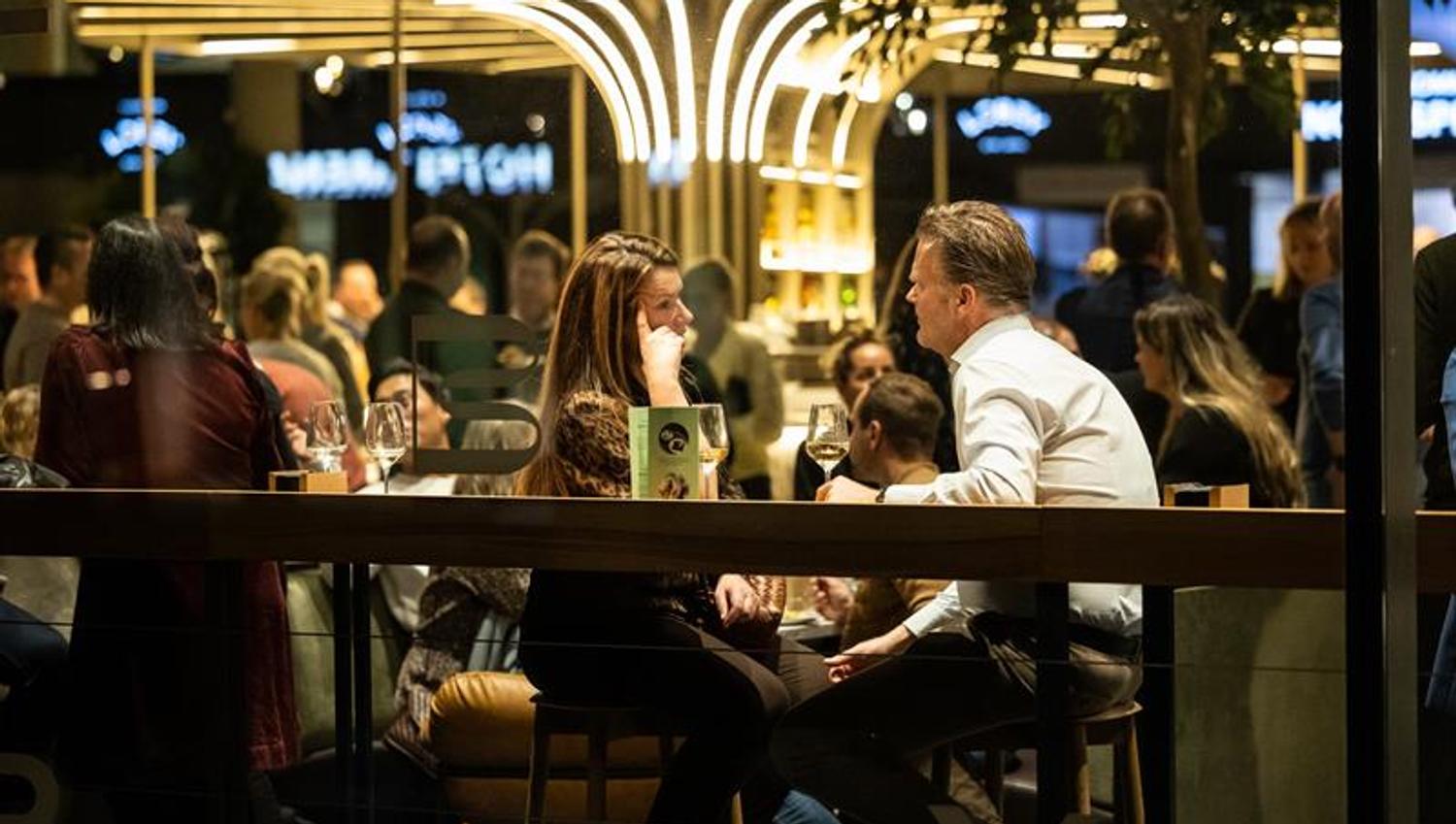En kvinne og en mann sitter ved et bord og snakker sammen over et glass vin i et travelt og stemningsfullt restaurant- eller barområde.