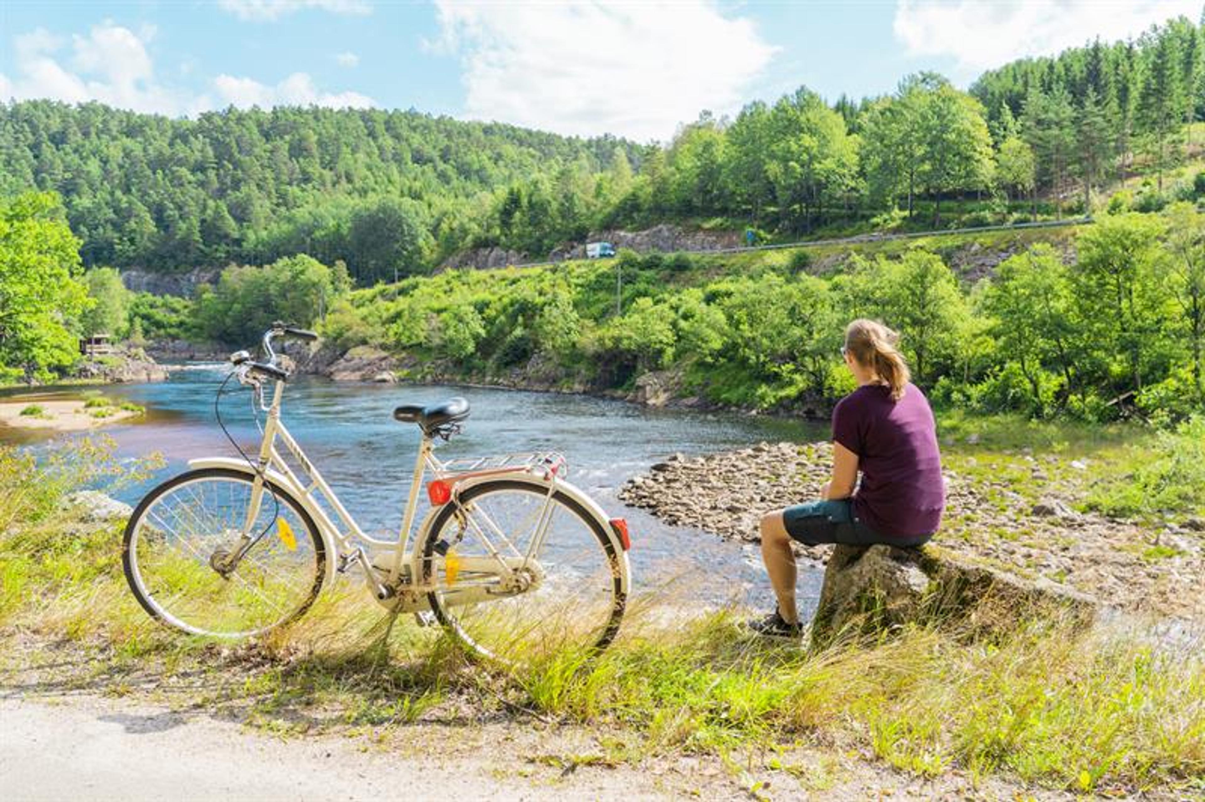 En dame sittende på en stein og ser mot Mandalselva, hvit sykkel vedsiden av henne.