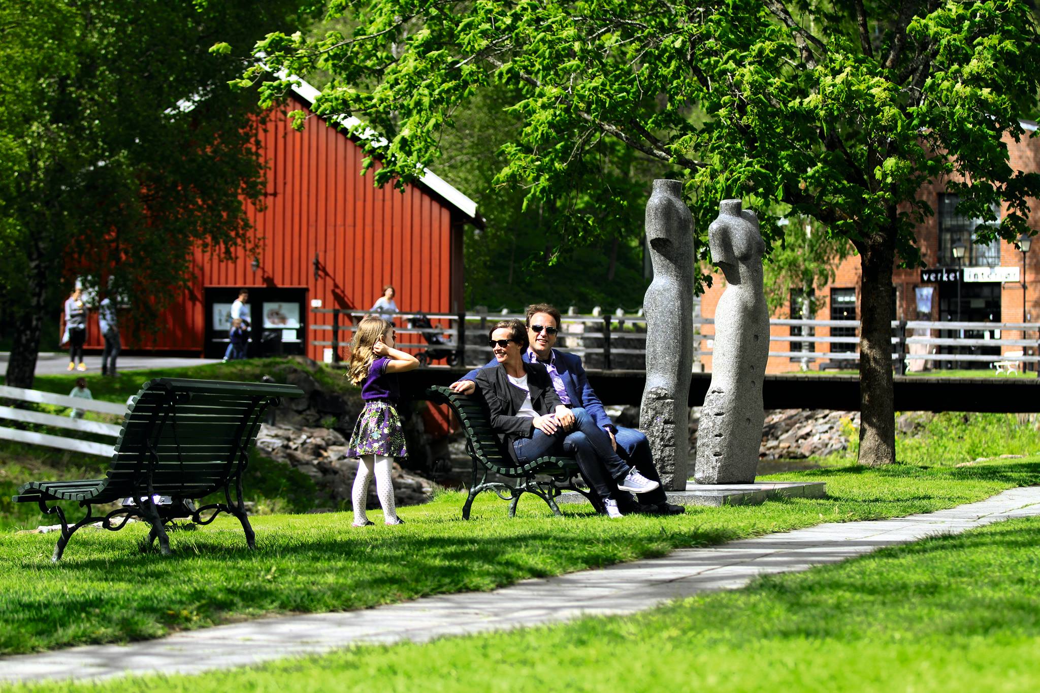 Bilde av familien som sitter på benk i solen med skulpturer rundt på det grønne gresset