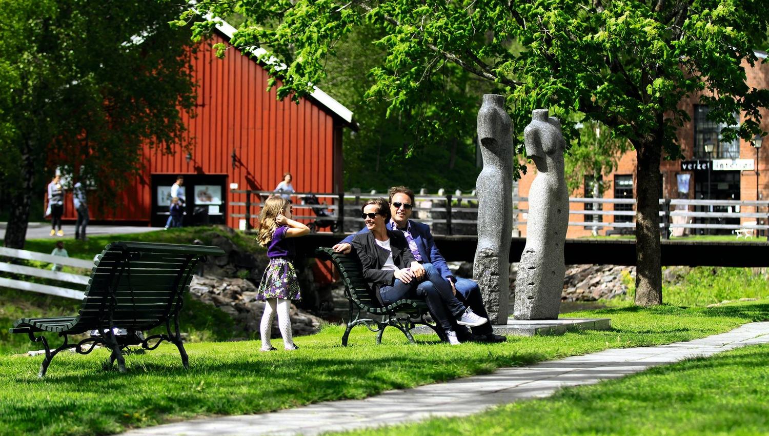 Bilde av familien som sitter på benk i solen med skulpturer rundt på det grønne gresset