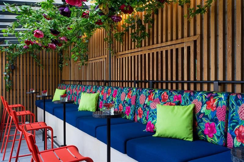 An outdoors seating area with floral upholstery and green cushions, small black round tables, and red chairs