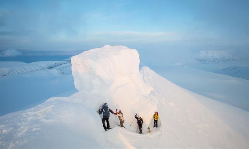 Fire personer som går langs en smal sti på toppen av et snødekt fjell, med fjellformasjonen kjent som Trollsteinen kledd i snø bak de