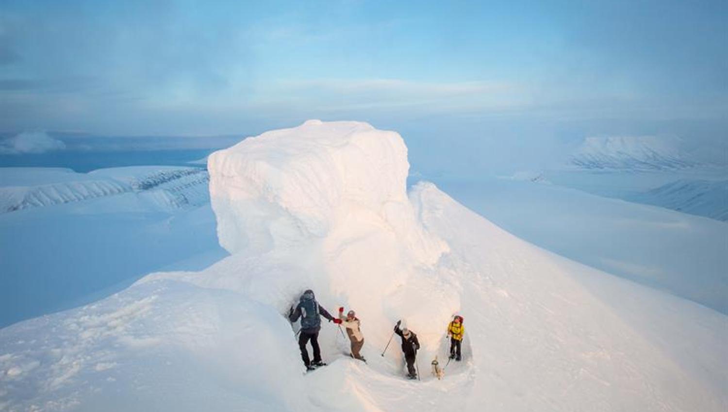 Fire personer som går langs en smal sti på toppen av et snødekt fjell, med fjellformasjonen kjent som Trollsteinen kledd i snø bak de