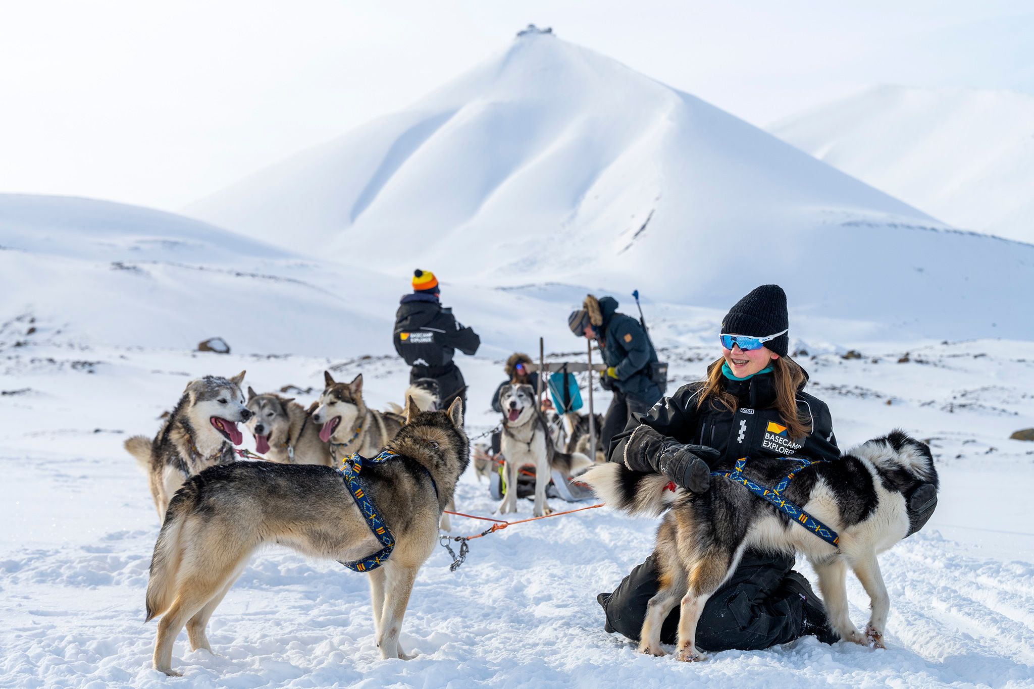 A guest cuddling with a dog in front of a sled dog team out in a snow-covered landscape. In the background a guide is standing on a sled and a guest a