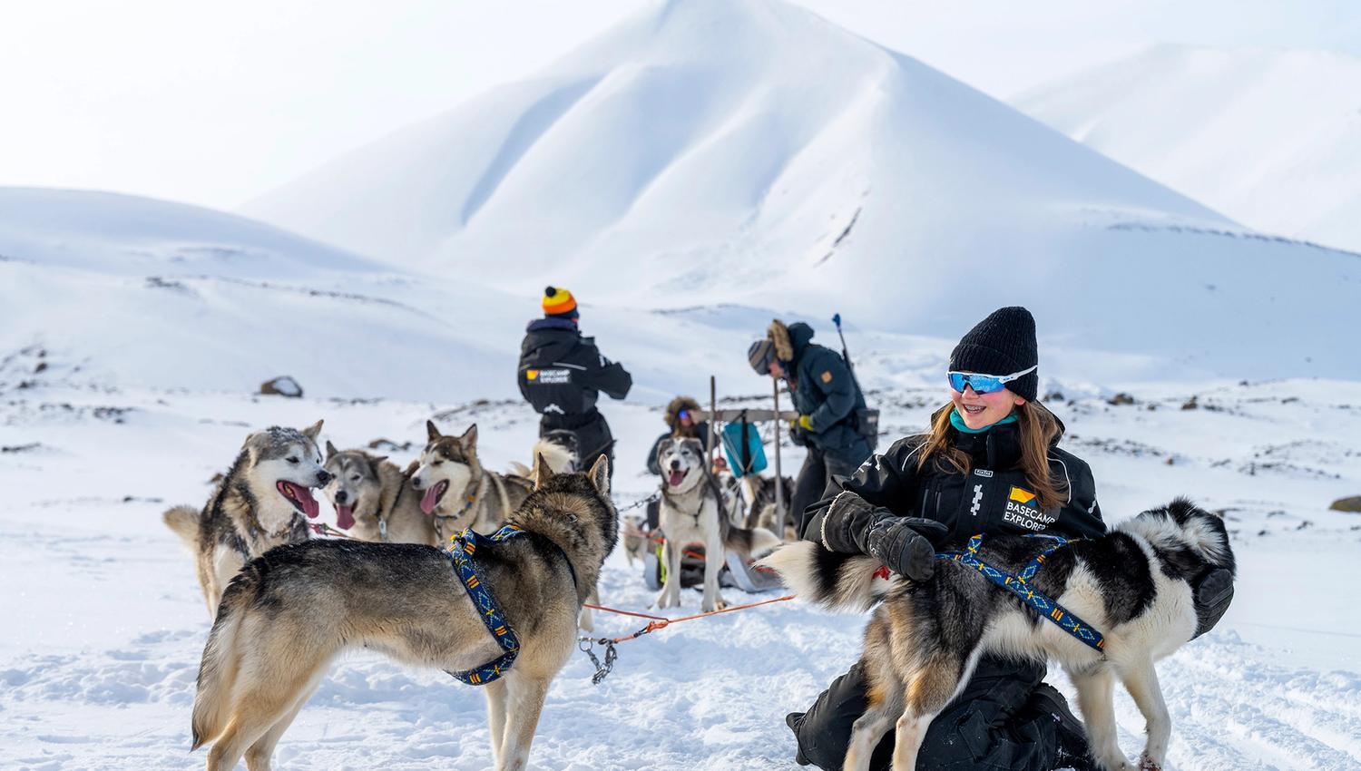 A guest cuddling with a dog in front of a sled dog team out in a snow-covered landscape. In the background a guide is standing on a sled and a guest a