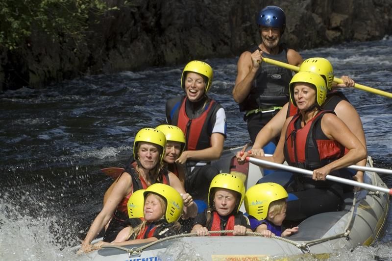 Rafting med mennesker i svarte våtdrakter og rød flytevest + gule hjelmer.