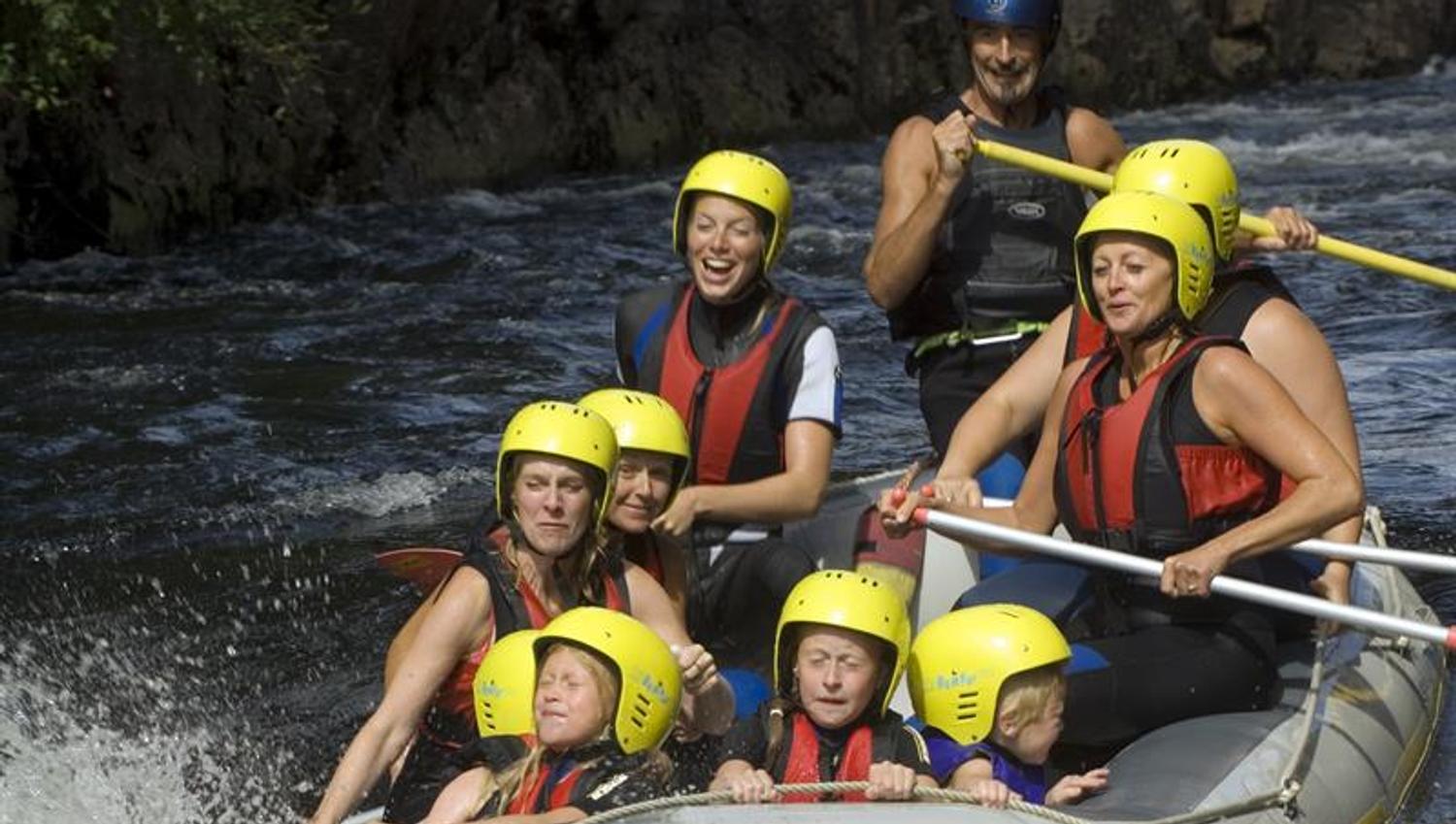 Rafting med mennesker i svarte våtdrakter og rød flytevest + gule hjelmer.