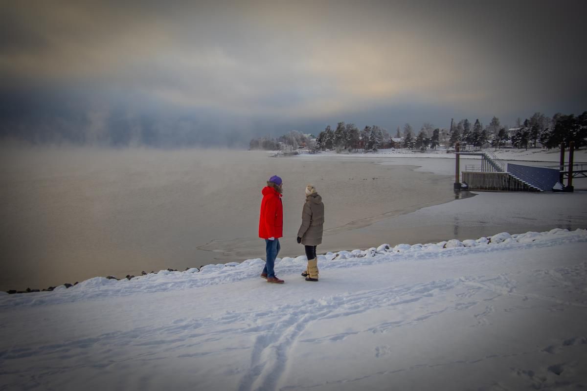To personer går på en snødekt sti langs et vann med tåke i bakgrunnen.
