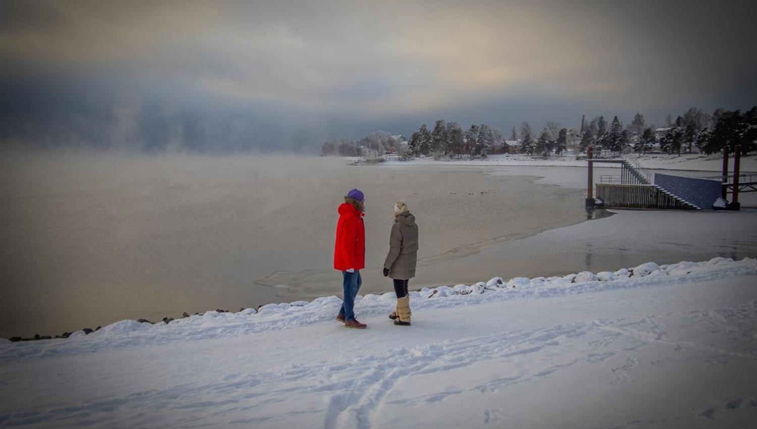 To personer går på en snødekt sti langs et vann med tåke i bakgrunnen.