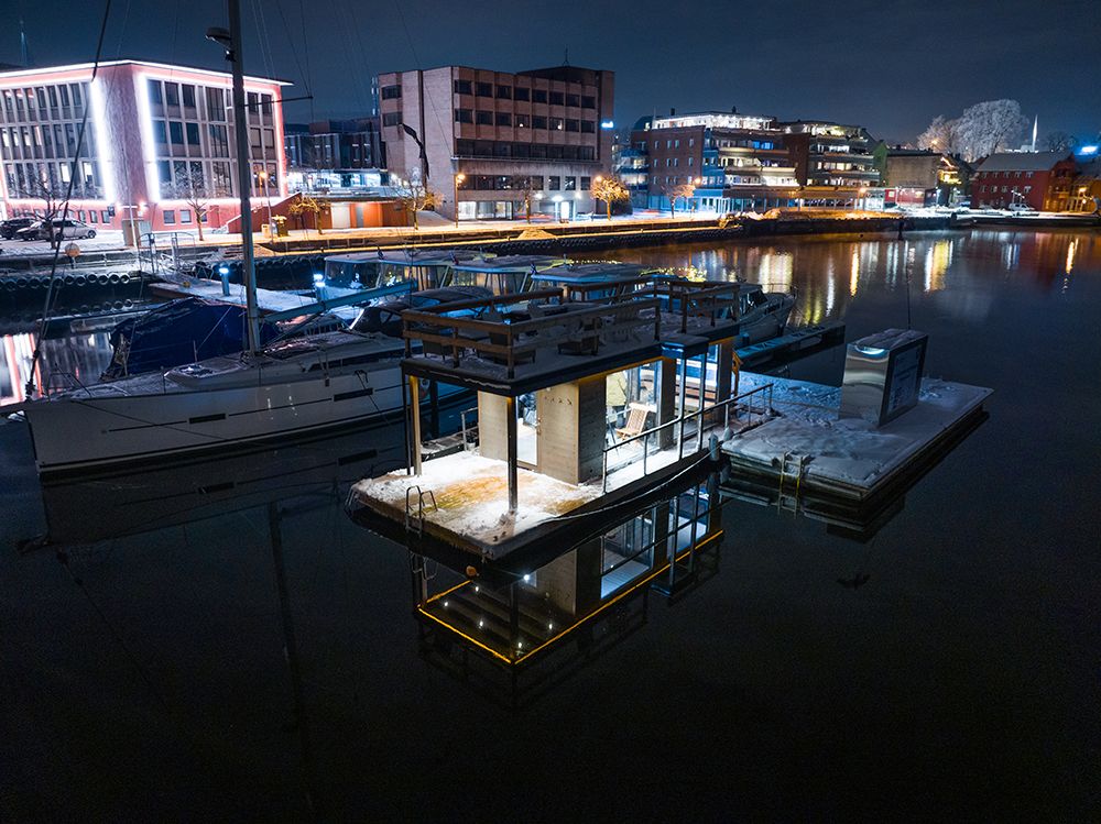 sauna liggene ved elva, med båter på brygge. blokker med lys i bakgrunnen