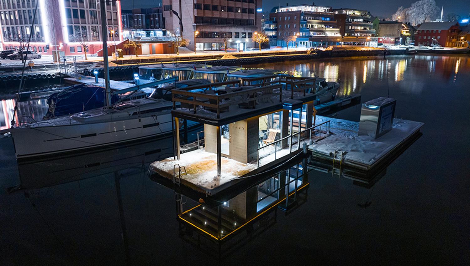 sauna liggene ved elva, med båter på brygge. blokker med lys i bakgrunnen