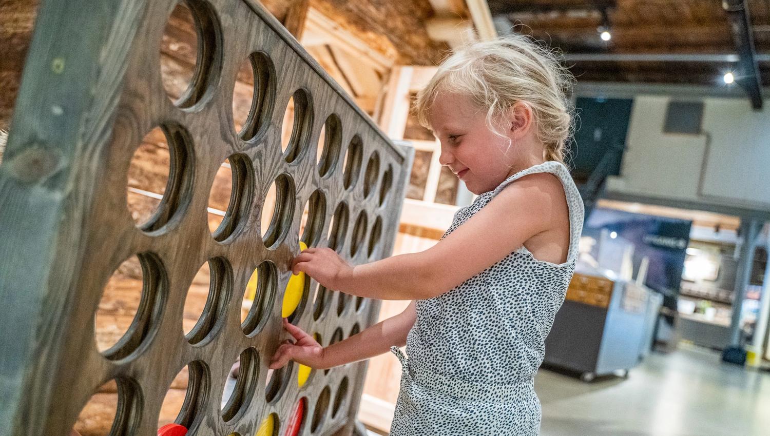 Bildet viser ei lita jente som spiller et stort fire på rad-spill laget av tre. Hun setter inn en rød brikke og smiler mens hun leker. Bak henne ser man deler av et museum med utstillinger.
