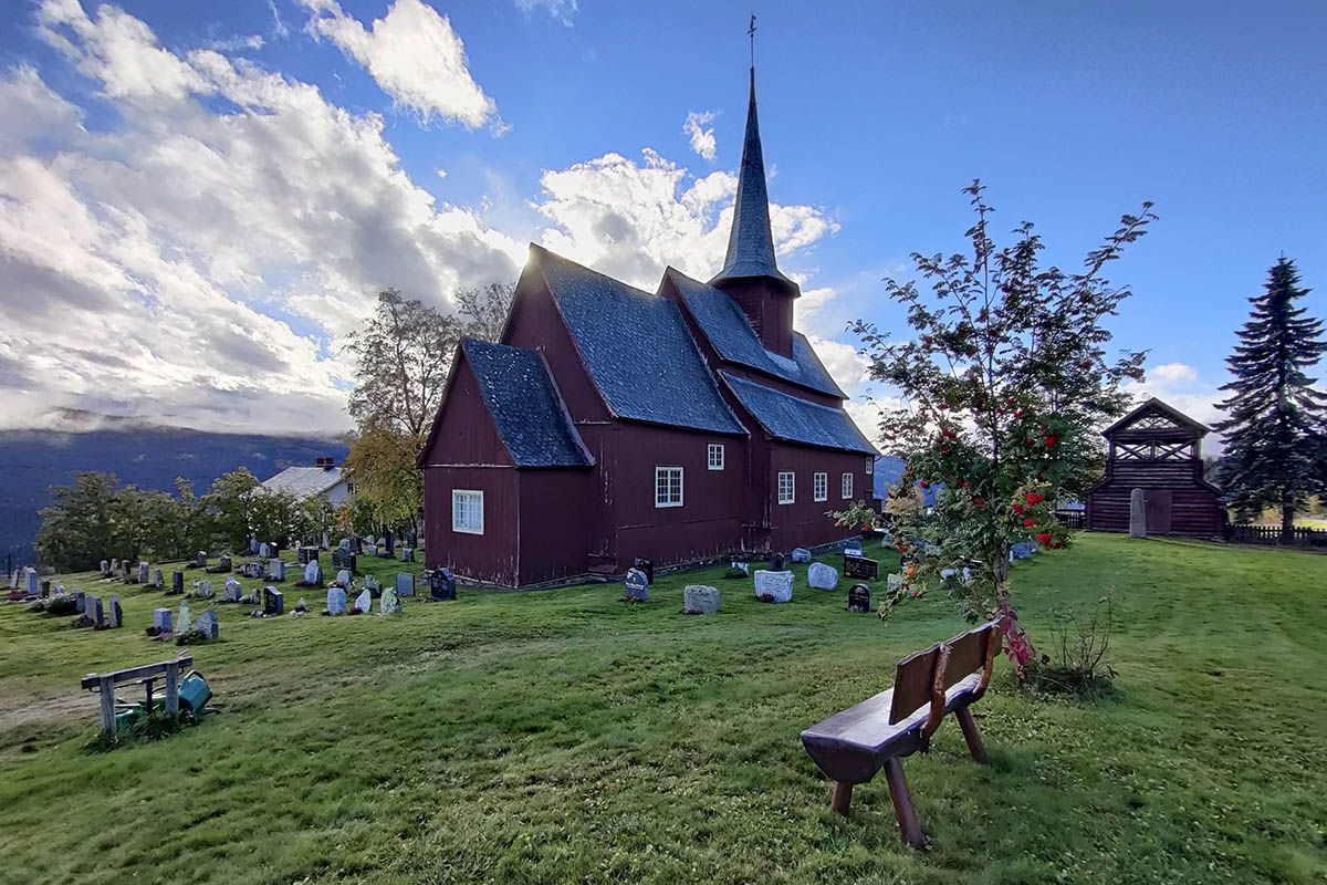 Stave church on a green lawn with a small rowan berry tree against the sun.