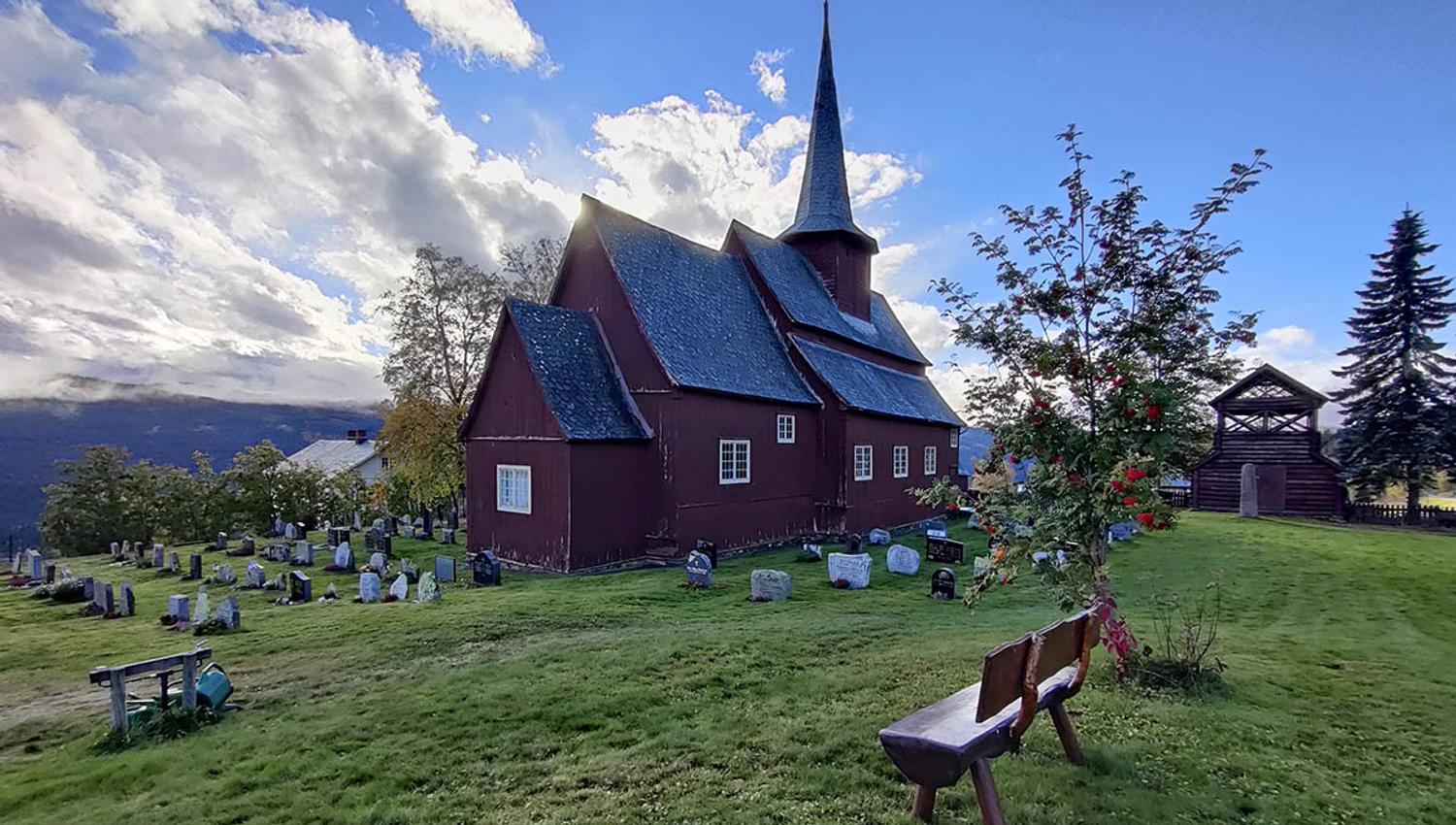 Stave church on a green lawn with a small rowan berry tree against the sun.