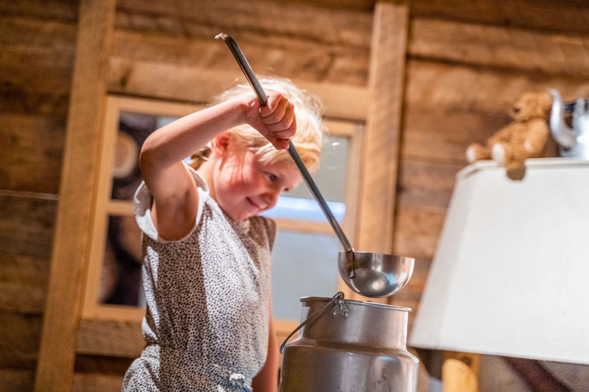 Bildet viser ei lita jente som holder en øse og later som hun heller noe fra en stor melkekanne. Hun smiler og ser ut til å leke inne i et rom med tømmervegger, kanskje et gammeldags museum.