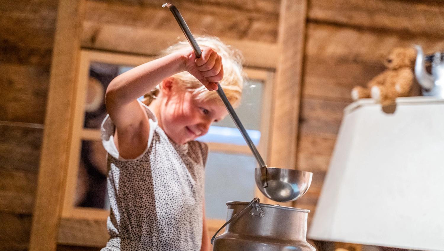 Bildet viser ei lita jente som holder en øse og later som hun heller noe fra en stor melkekanne. Hun smiler og ser ut til å leke inne i et rom med tømmervegger, kanskje et gammeldags museum.