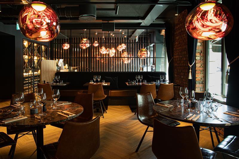 Restaurantinteriør med runde, brune bord, brune stoler og sitteseksjon mot veggen.