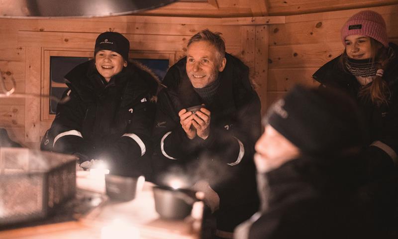 Personer sitter i en koie rundt åpen ild